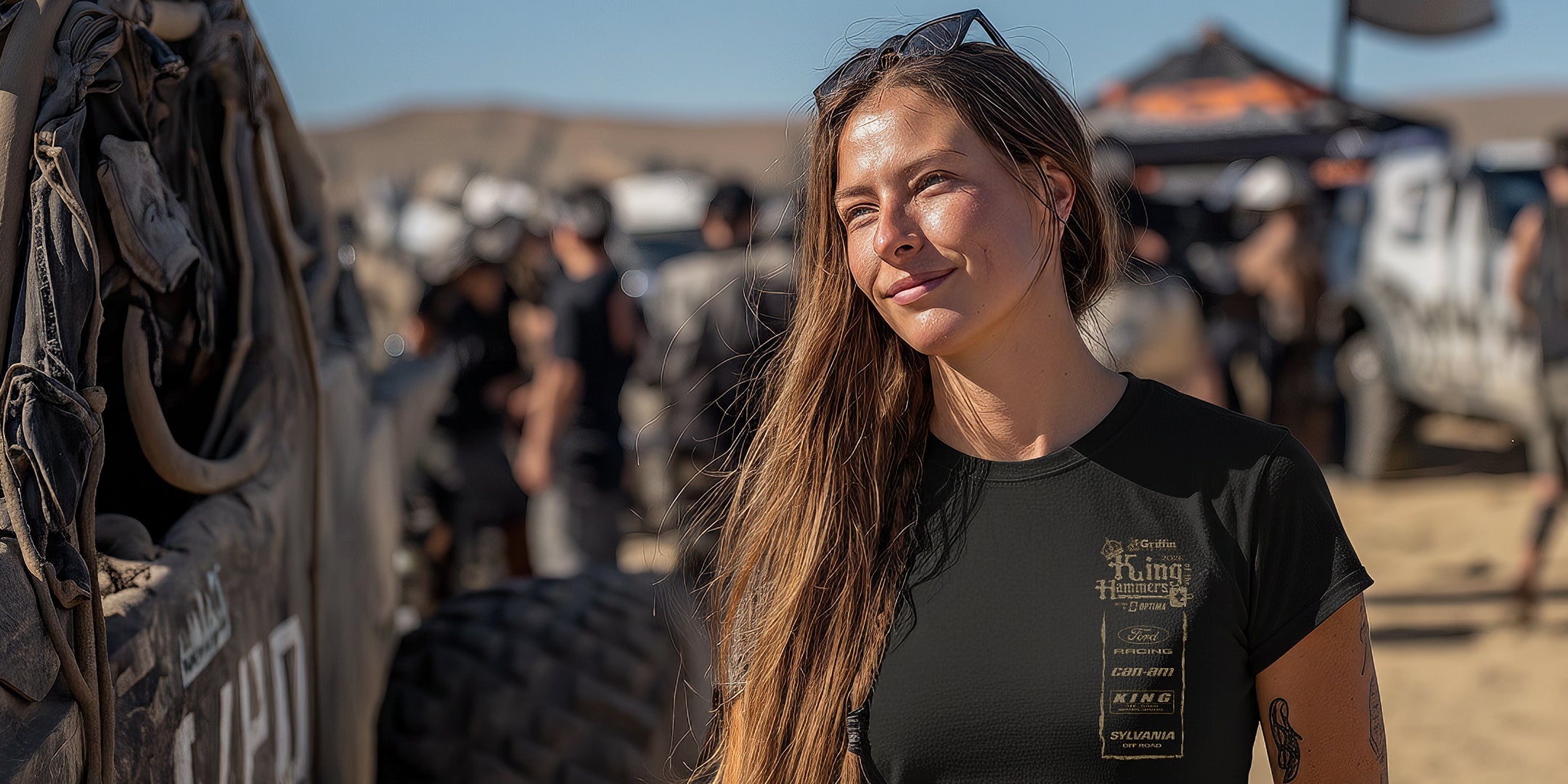 Woman in a black King of the Hammers t-shirt standing in a desert-like setting with vehicles and people in the background