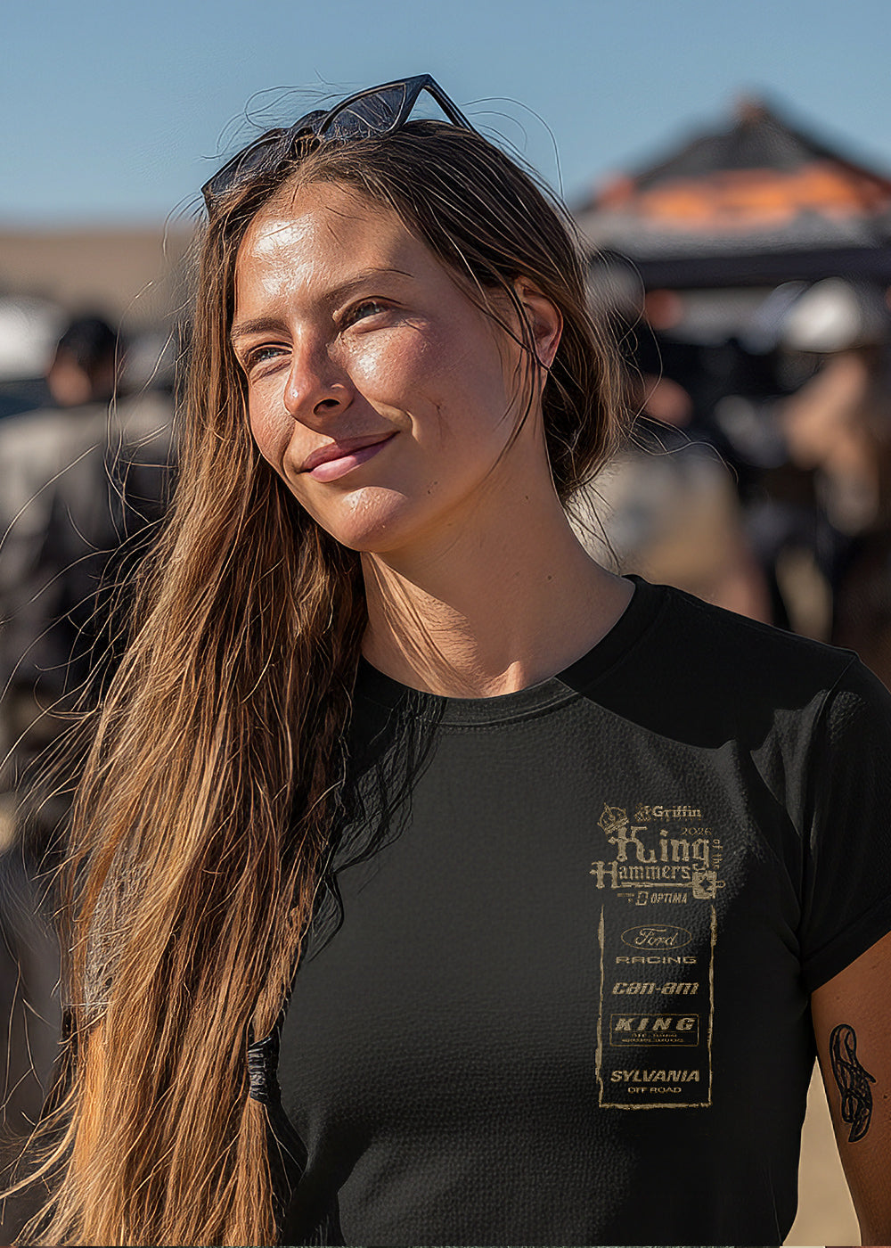 Woman in a black King of the Hammers t-shirt standing in a desert-like setting with vehicles and people in the background