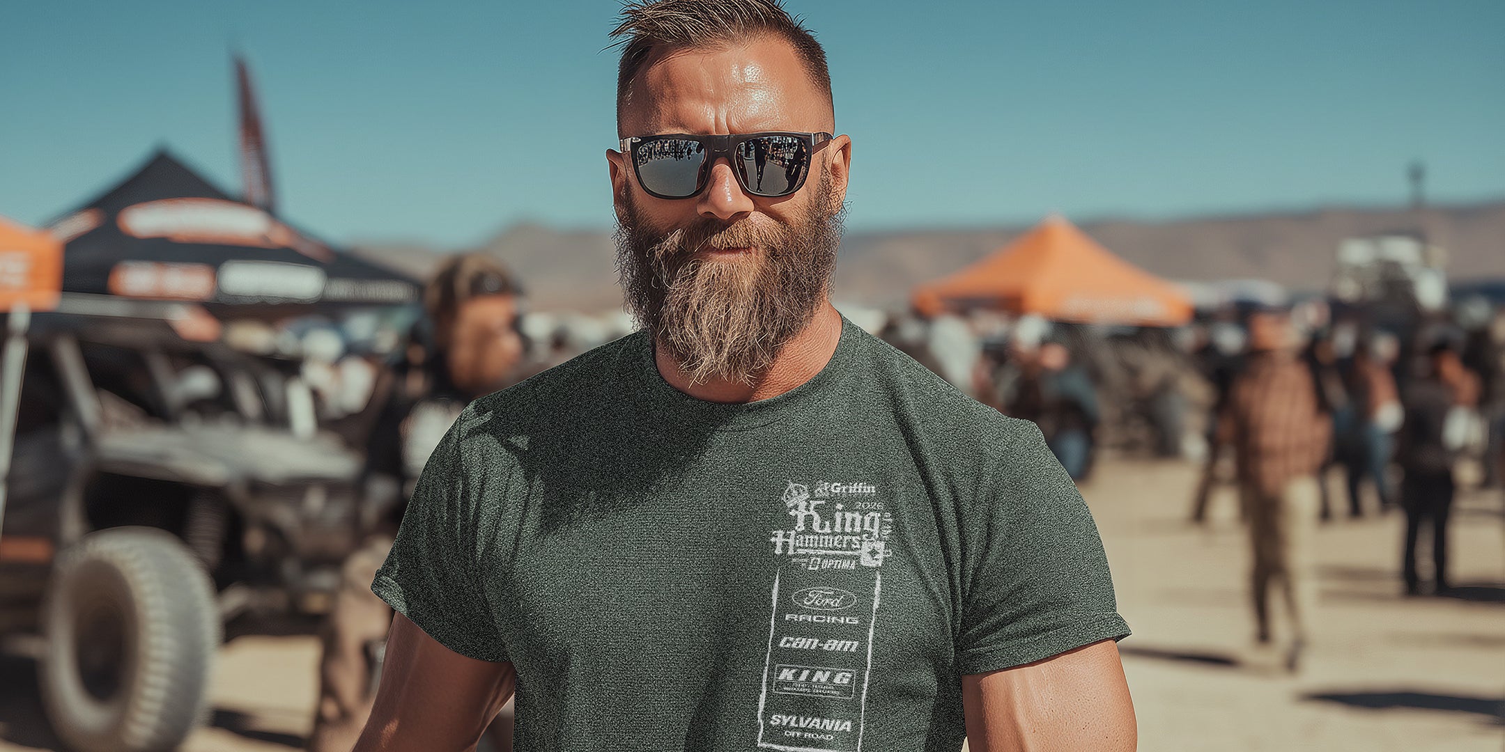 Man with sunglasses and beard standing in a desert event with vehicles and tents in the background