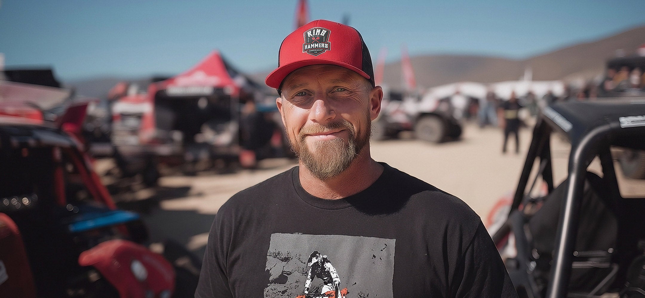 Man wearing a red cap with a King of the Hammers logo and black t-shirt with a King of the Motos design, standing in an outdoor setting with vehicles and tents.
