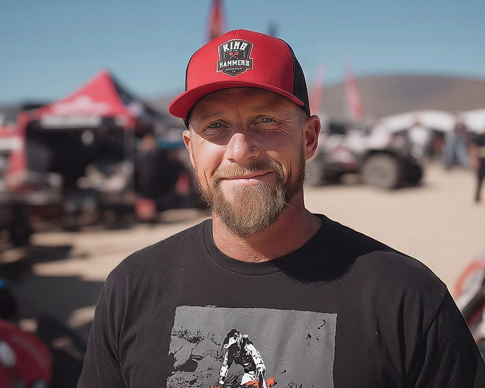 Man wearing a red cap with a King of the Hammers logo and black t-shirt with a King of the Motos design, standing in an outdoor setting with vehicles and tents.