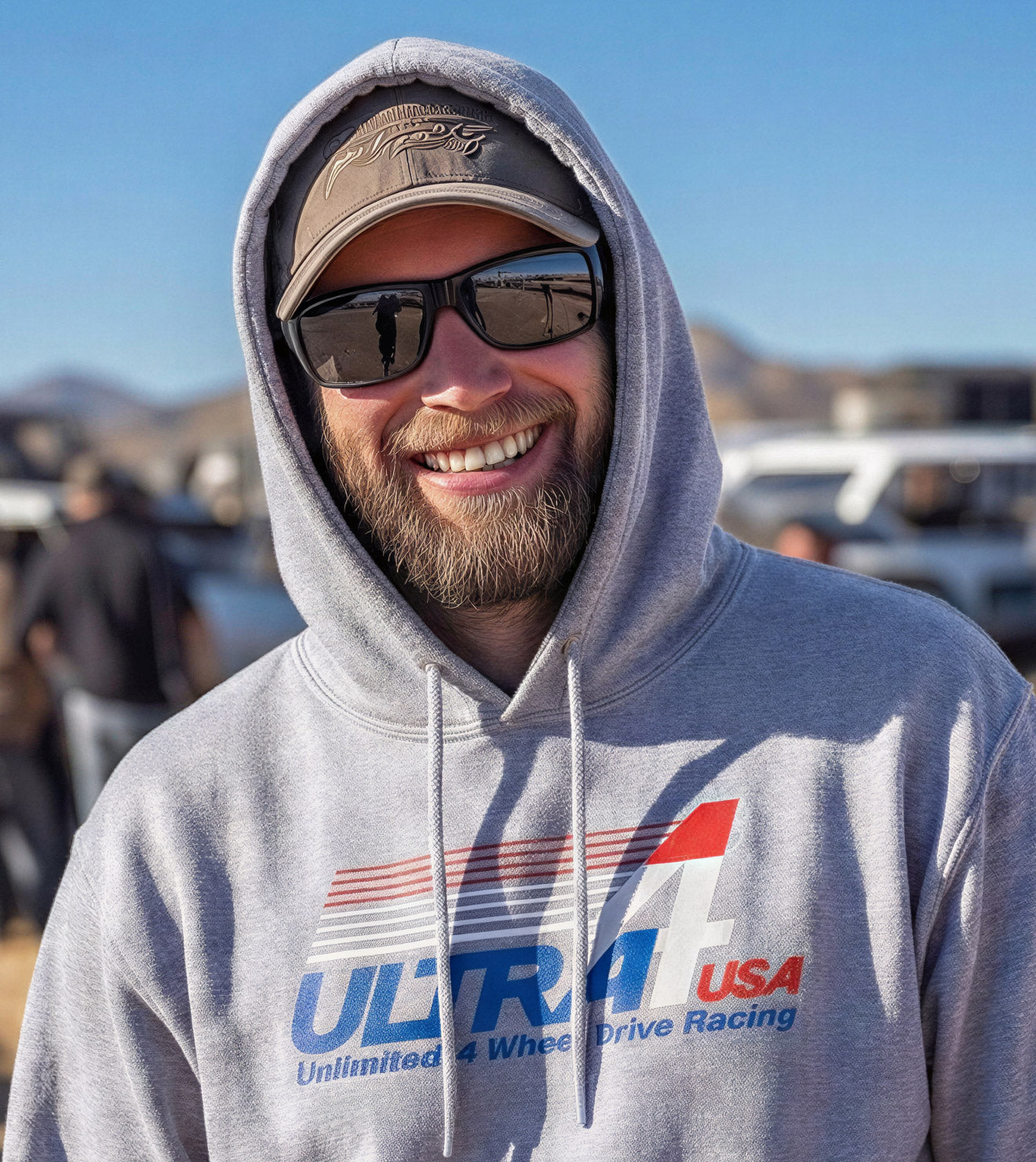 Man wearing a gray hoodie with 'Ultra 4 USA' logo, sunglasses, and cap in an outdoor setting.