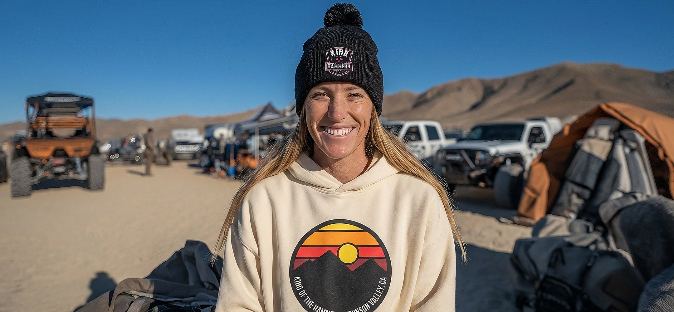 Smiling woman wearing a beige hoodie with a King of the Hammers desert logo and black beanie with a King of the Hammers logo, standing in a desert with off-road vehicles and mountains in the background.