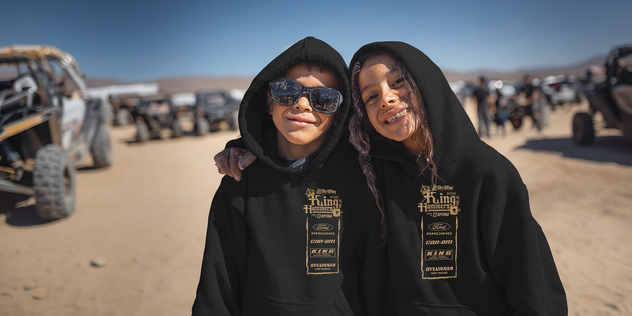 Two kids wearing black hoodies with King of the Hammers branding in a desert setting with off-road vehicles.