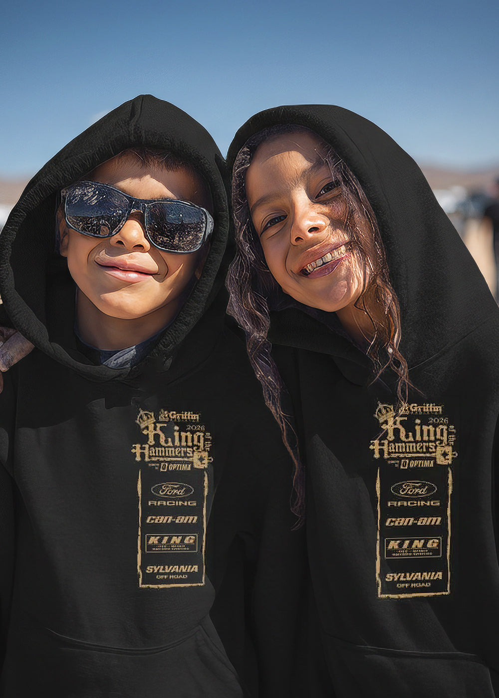 Two kids wearing black hoodies with King of the Hammers branding in a desert setting with off-road vehicles.