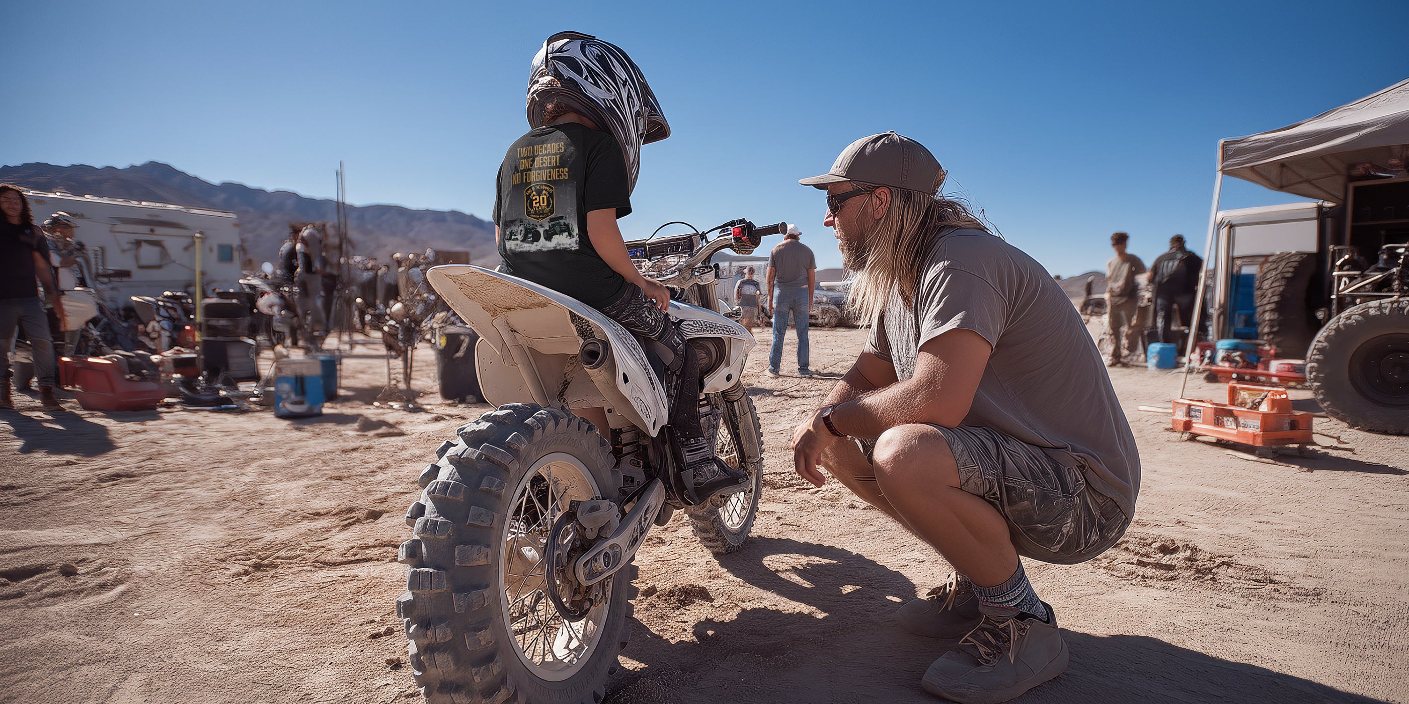 Father with his son wearing a King of the Hammers t-shirt and motorcycle helmet sitting on a dirt bike in an outdoor setting with vehicles and people in the background.