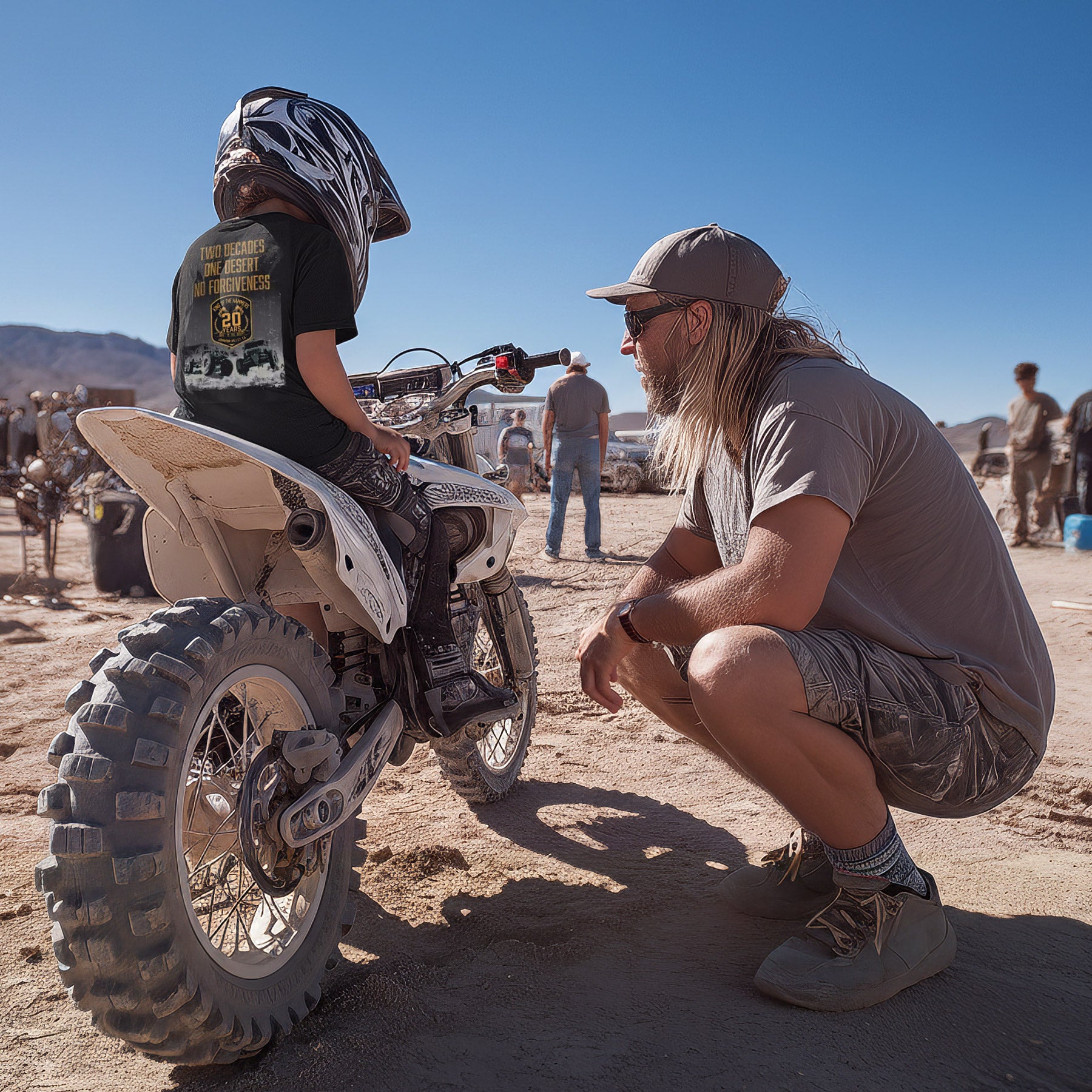 Father with his son wearing a King of the Hammers t-shirt and motorcycle helmet sitting on a dirt bike in an outdoor setting with vehicles and people in the background.