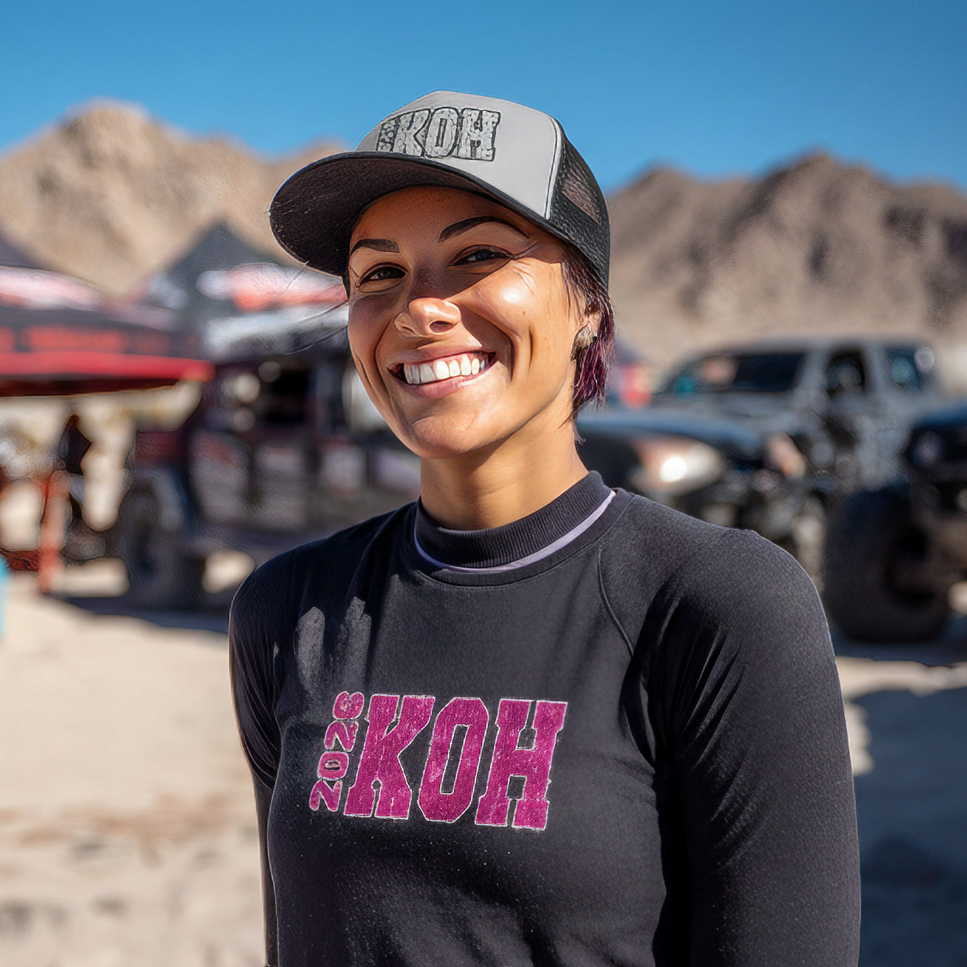 Smiling woman wearing a black shirt with '2020 KOH' text and a cap in a desert setting with vehicles and mountains.