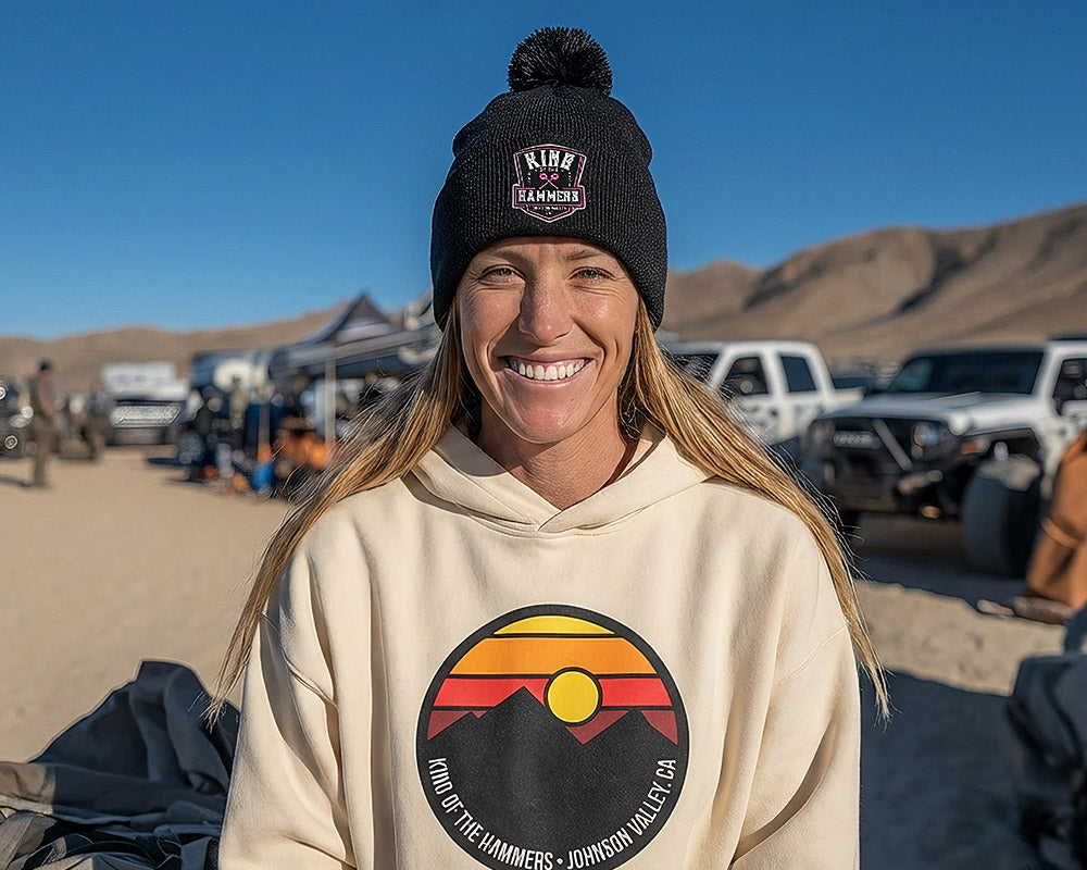 Smiling woman wearing a beige hoodie with a King of the Hammers desert logo and black beanie with a King of the Hammers logo, standing in a desert with off-road vehicles and mountains in the background.